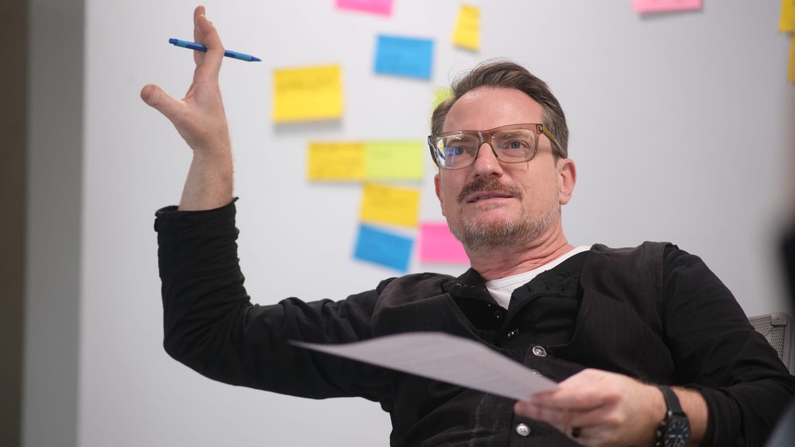 Manuel Kreutz erklärt mit erhobener Hand und einem Blatt Papier vor einer Wand mit bunten Haftnotizen